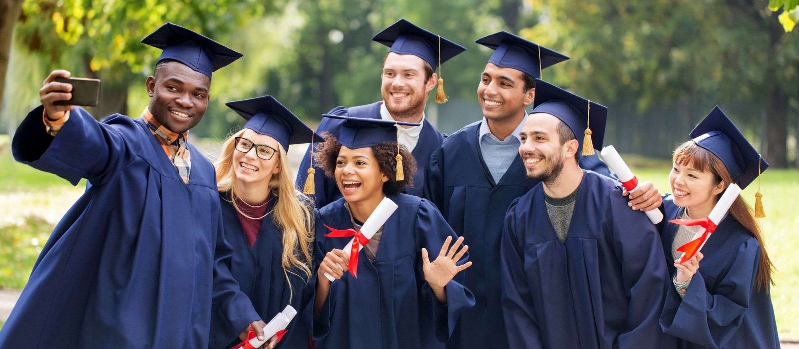 Group of diverse graduates in blue caps and gowns smiling and taking a selfie outdoors while holding diplomas
