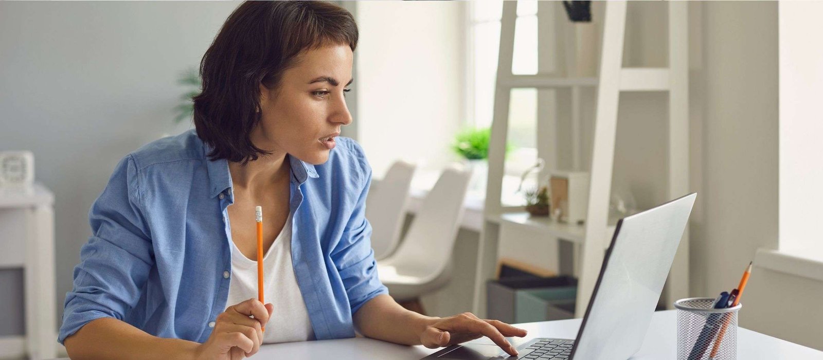 Young woman holding a pencil and looking intently at her laptop screen while participating in Italian lessons online in a bright, modern workspace