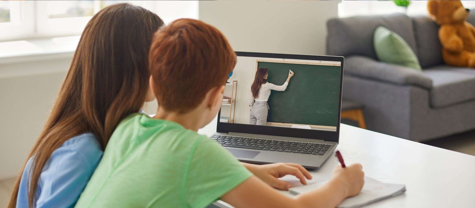 Two children sitting at a desk, participating in online German classes, watching a teacher on the laptop screen as one child writes notes in a notebook