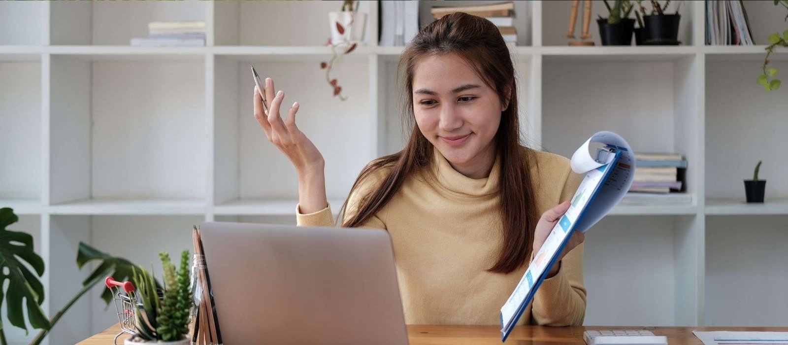 Smiling woman sitting at a desk, engaging in an online Russian course while holding a folder and talking to her laptop in a bright home office